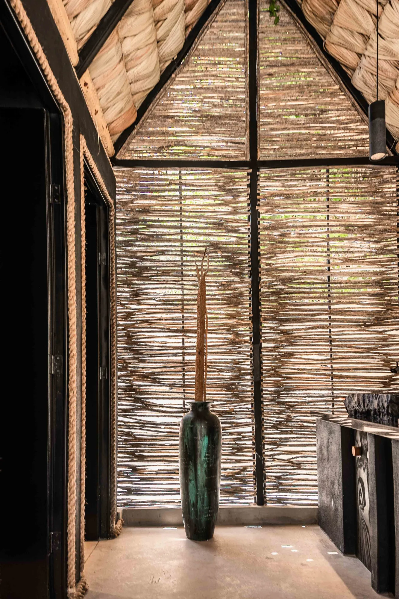 Restroom vignette of a green-glazed jar with a dried palm stalk against an ocotillo-stick wall by a rope curtain