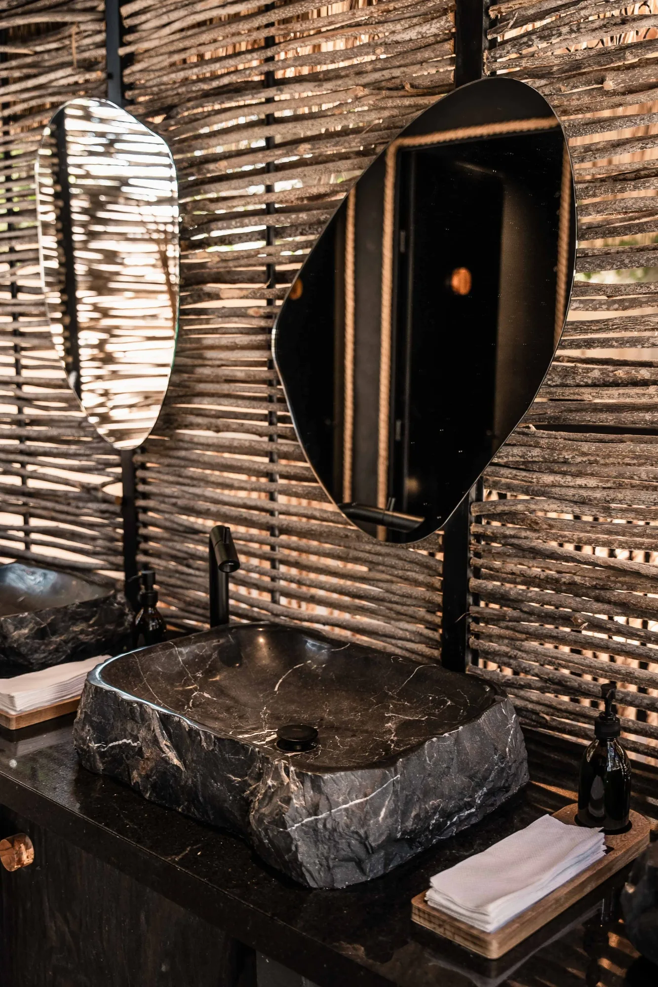 Vanity detail with a rough black stone vessel sink, organic mirrors and a black faucet against a stick wall