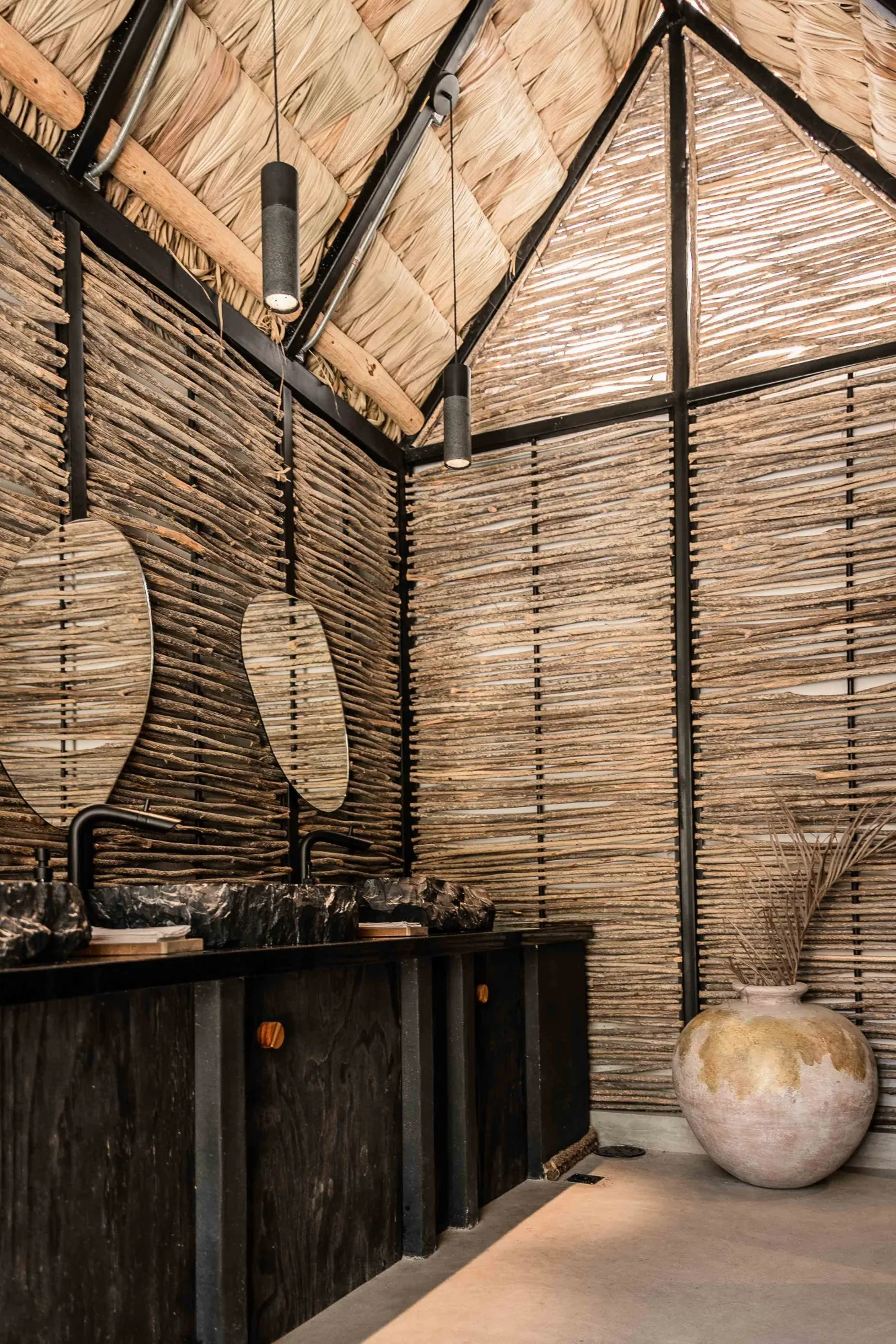 Restroom with two stone vessel sinks, leaf-shaped mirrors and cone pendants under a thatched roof and ocotillo-stick walls