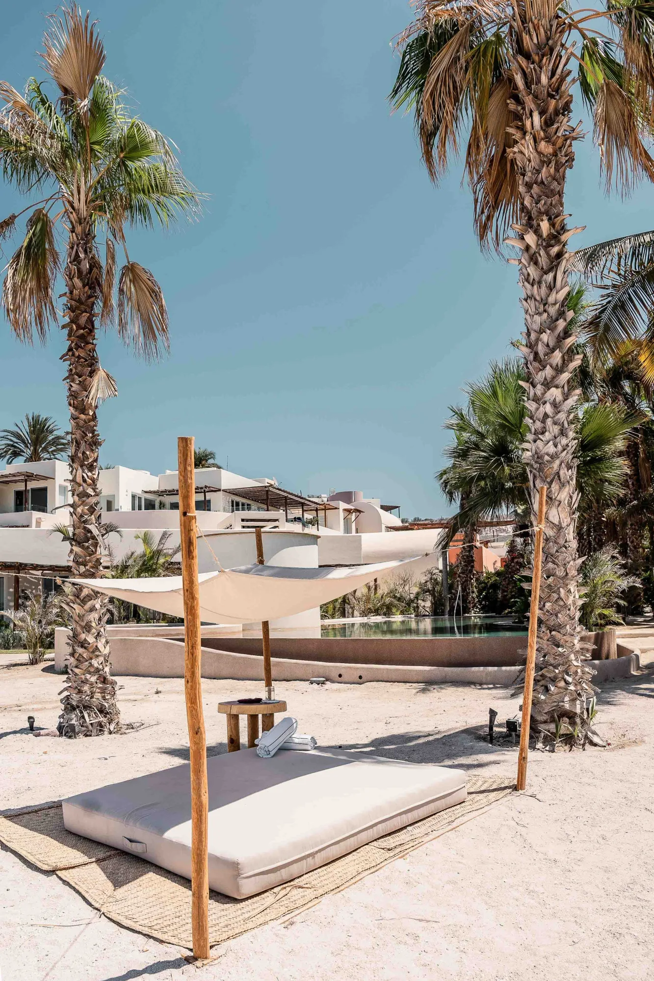 Beach daybed under a canvas shade sail on bamboo poles between two tall palms, the pool and white buildings behind