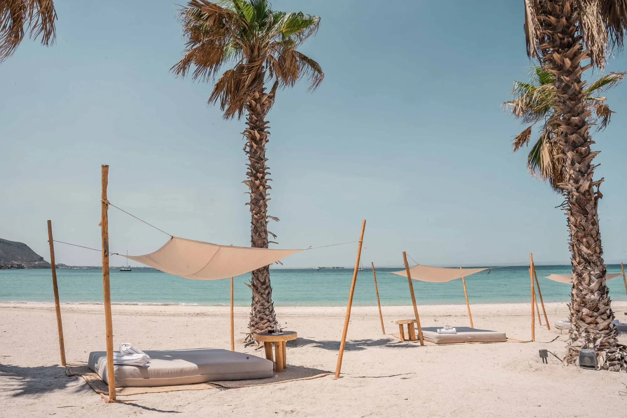 Canvas shade sails on bamboo poles over beach daybeds between palms, the turquoise Sea of Cortez behind