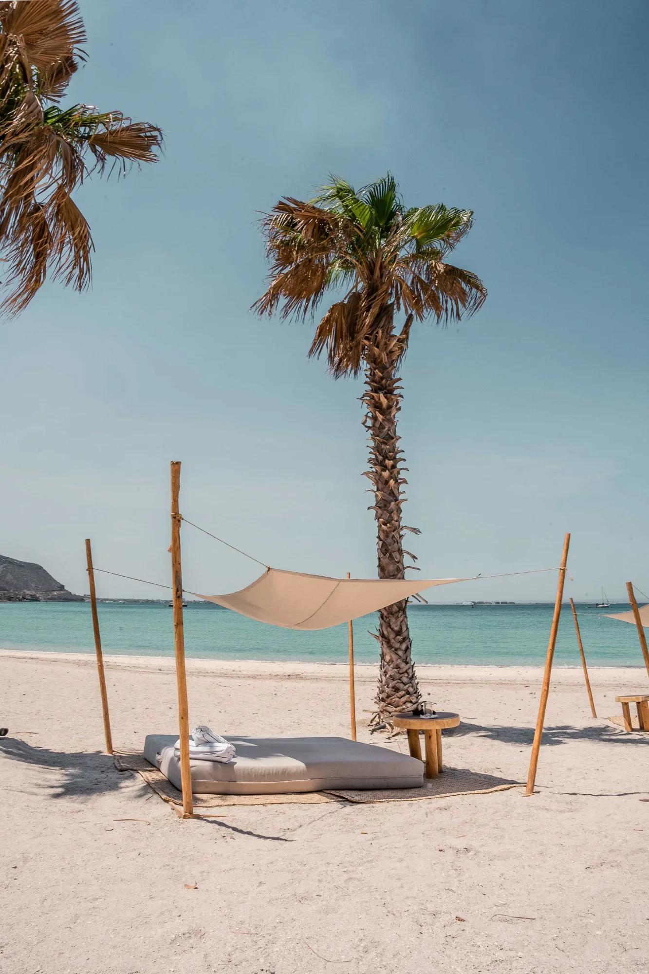 A single beach daybed under a canvas sail by a palm, the turquoise bay and a distant sailboat beyond