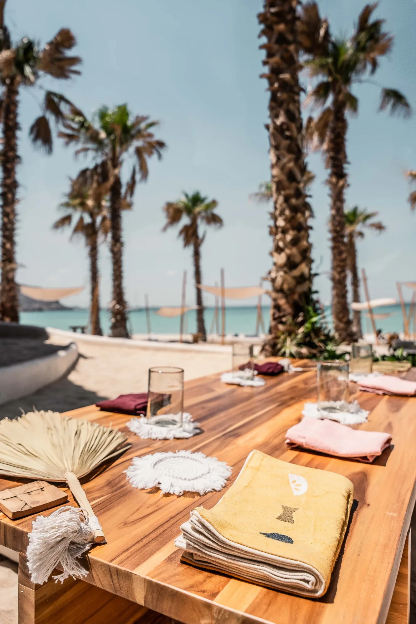 Communal teak table set with candle holders and printed napkins, palms and the bay with shade sails beyond