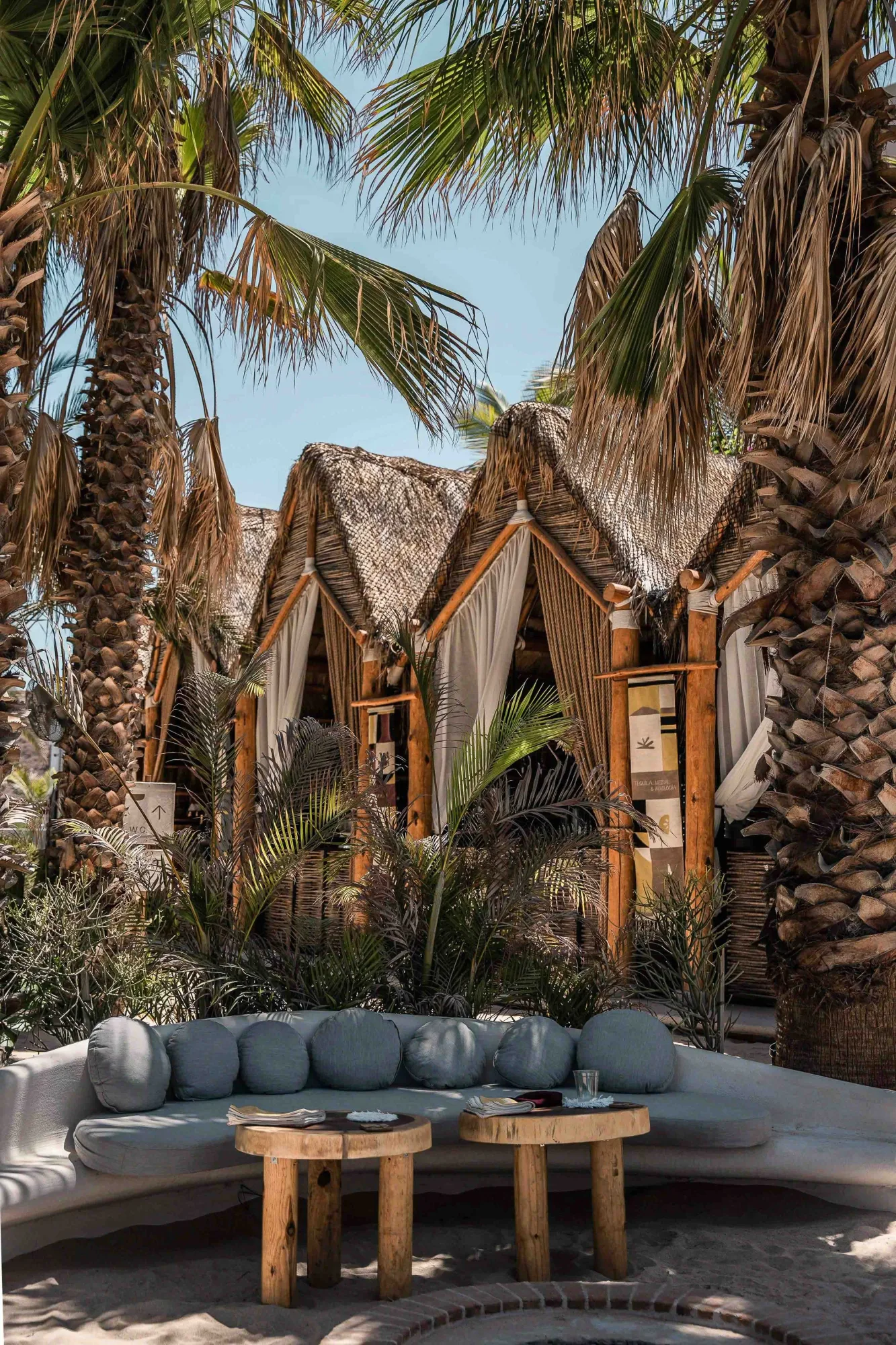 Curved plaster sofa with blue cushions and round wood tables facing palapa cabanas among lush palms