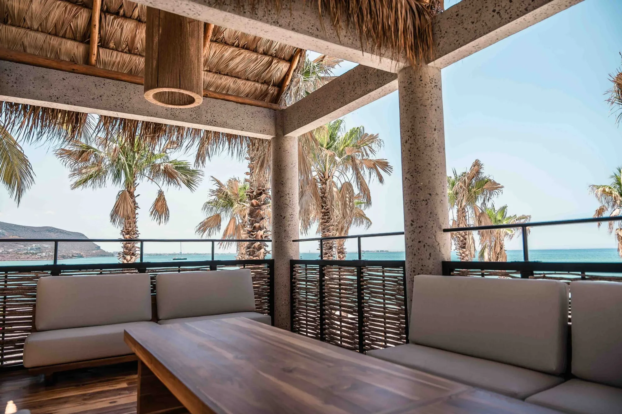 Covered upper terrace under a palapa on stone columns, grey sofas and a teak table open to palms and the Sea of Cortez