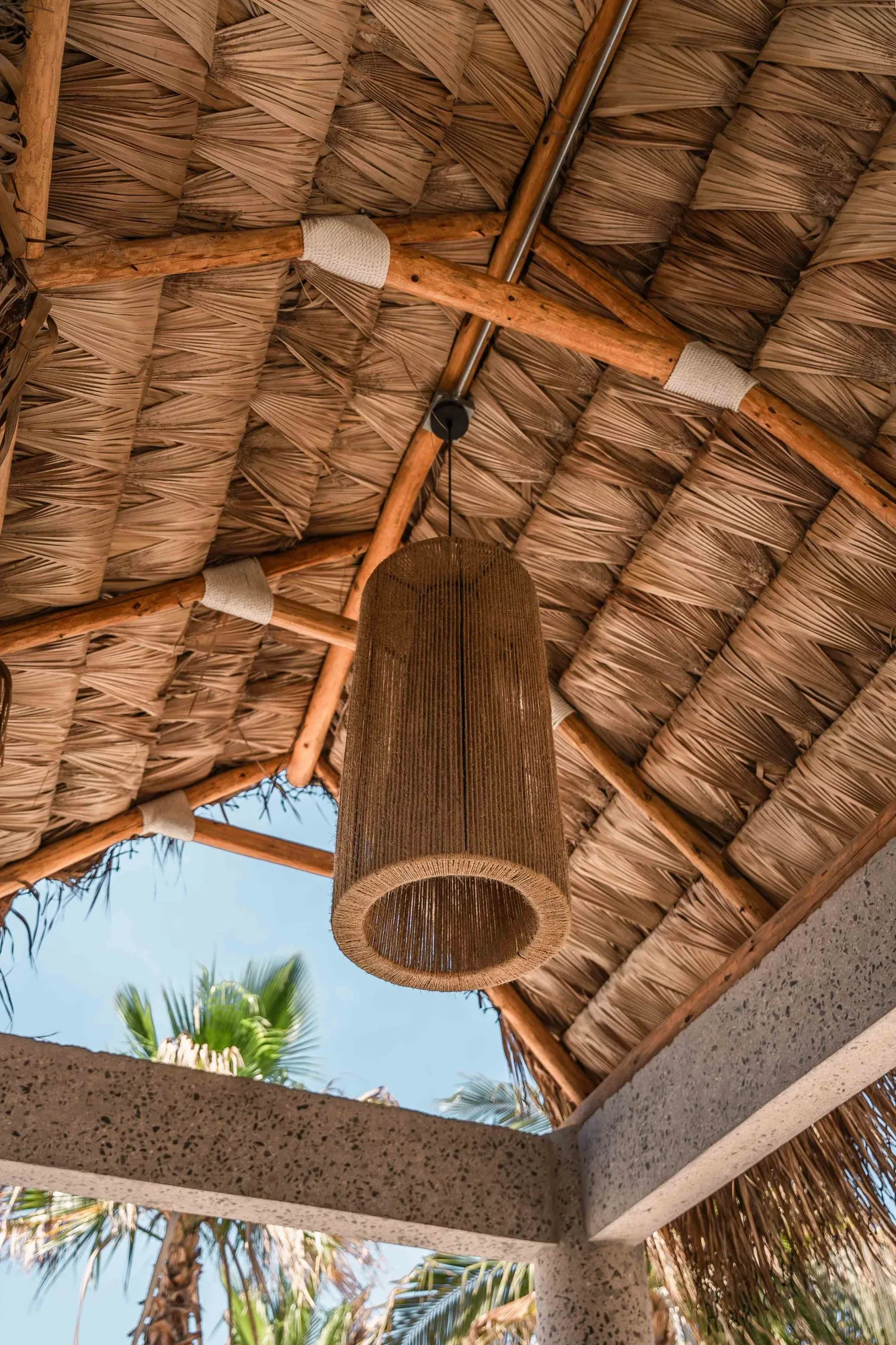 Cylindrical woven rattan pendant hanging from a palm-thatch ceiling on timber beams, palms and sky below