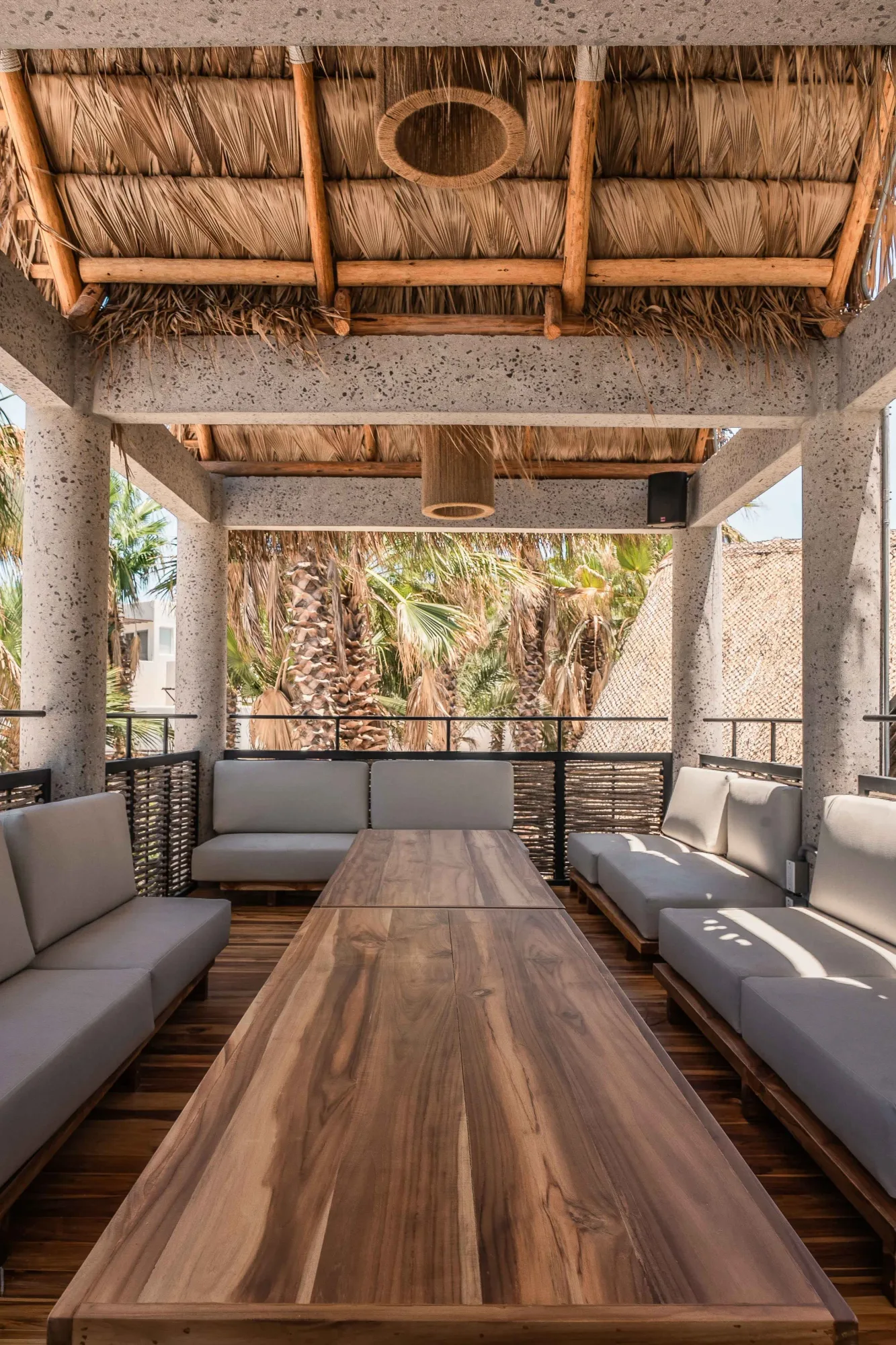 Covered terrace under a palapa with a woven pendant, grey sofas flanking a long teak table and woven railings