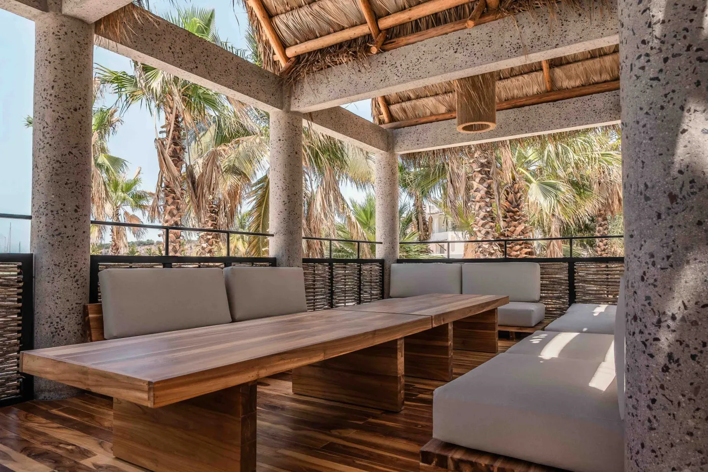Palapa-covered terrace with grey sofas around a teak table on stone columns, palm views all around