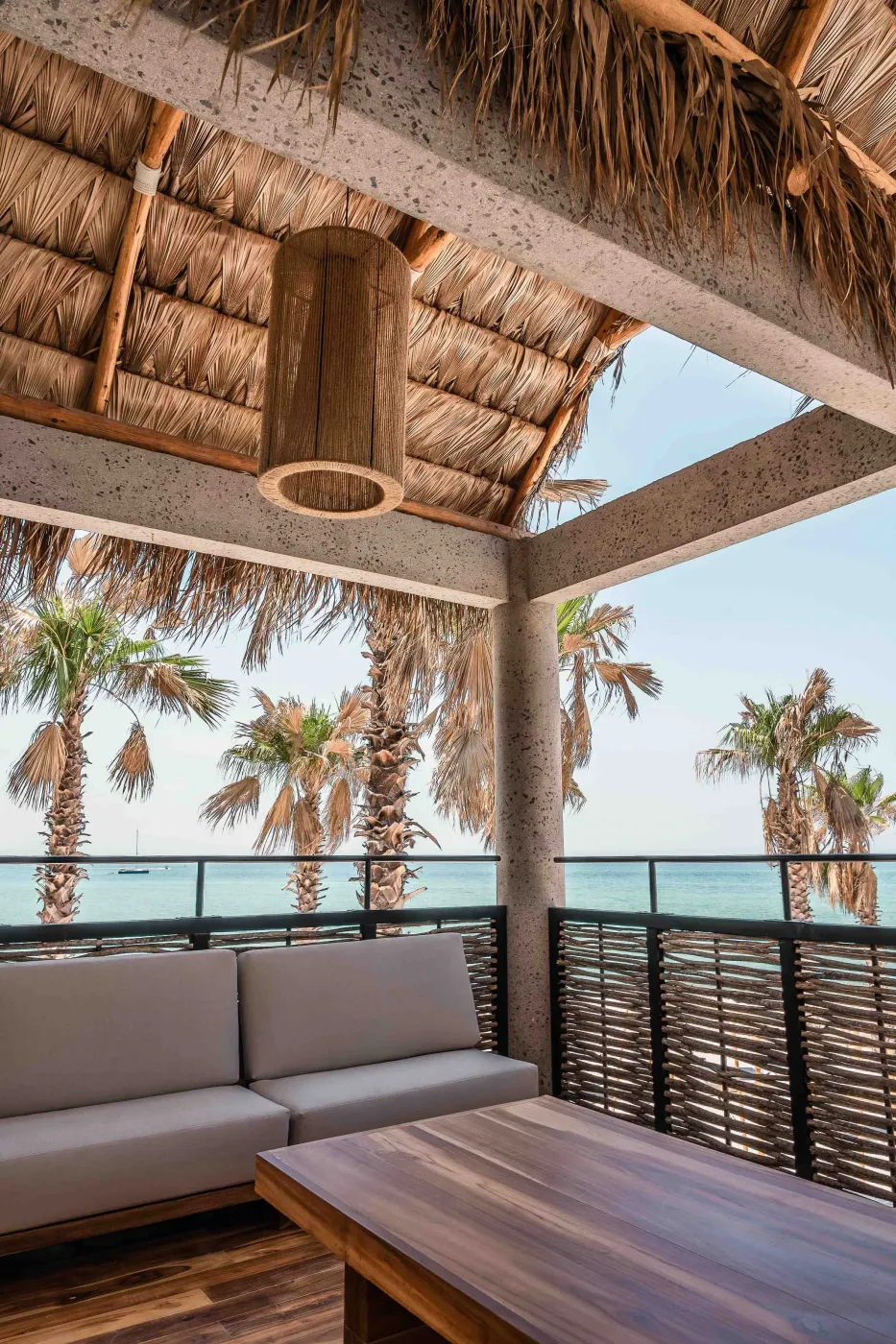 Terrace corner under a palapa with a woven pendant, grey sofa and teak table open to palms and the turquoise Sea of Cortez