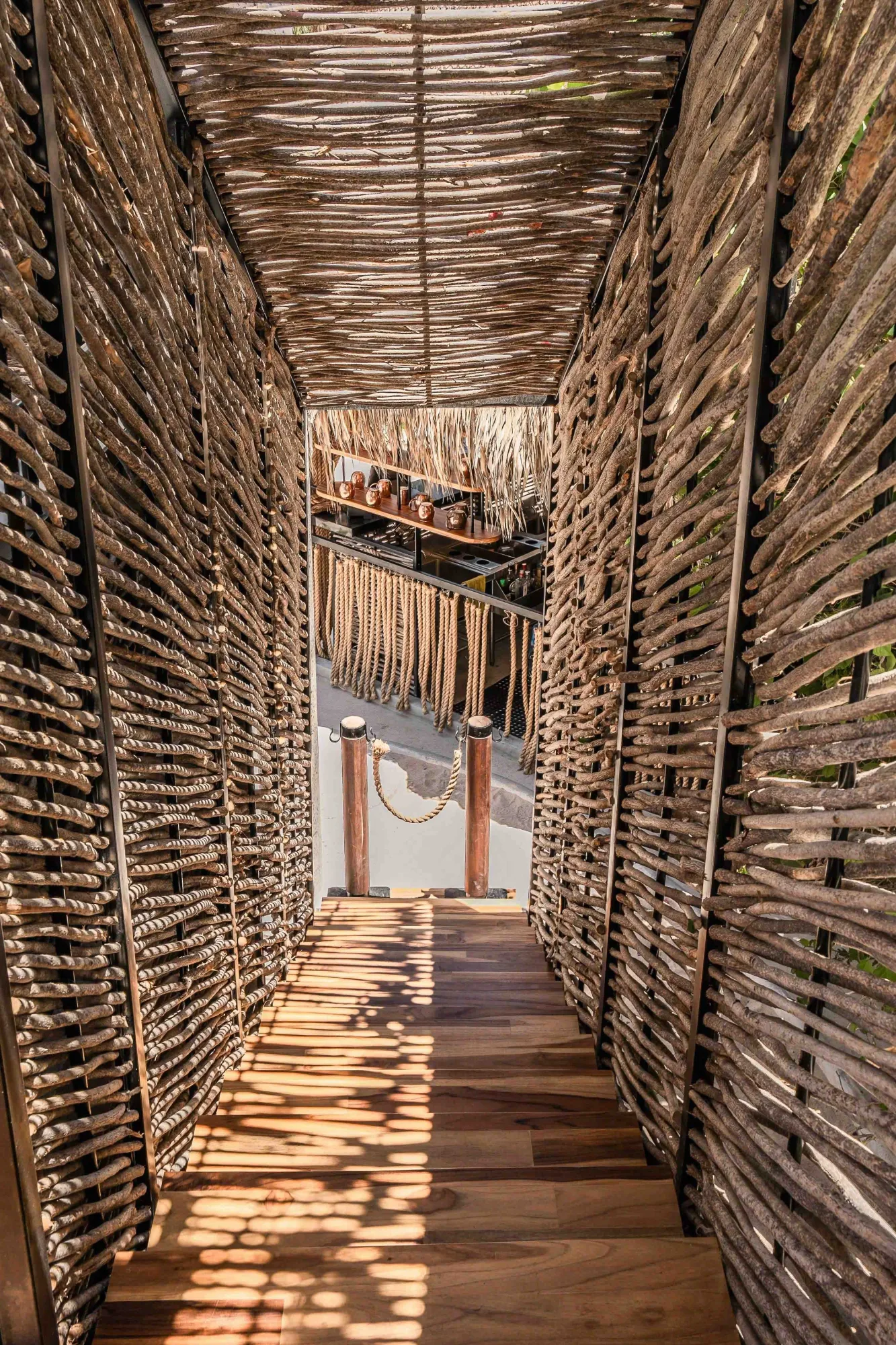 Corridor between tall ocotillo-stick walls with a wood-plank floor dappled with light toward a rope-fringe threshold