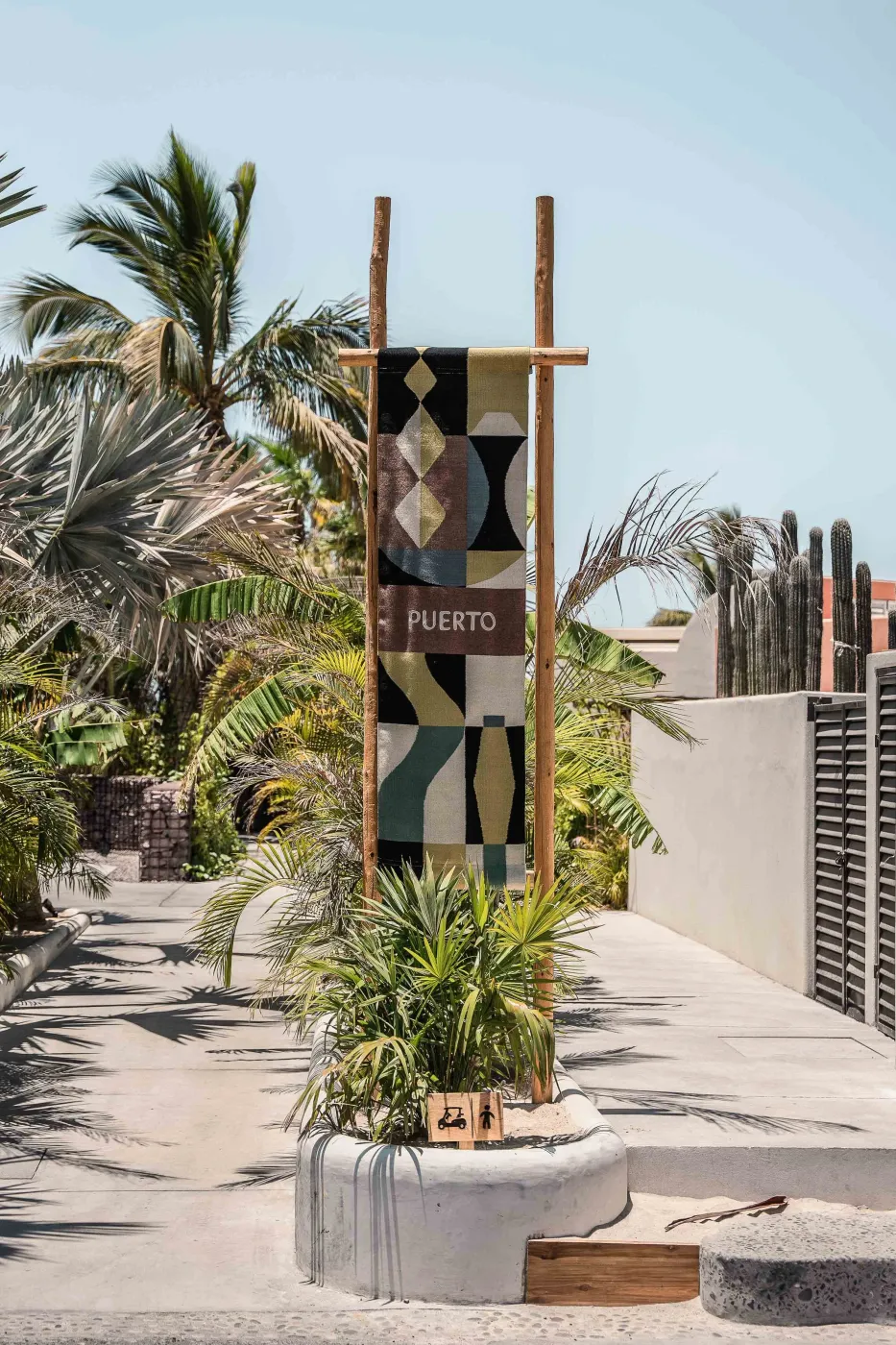 A graphic Puerto banner on a timber frame above a concrete planter with a palm along the walkway