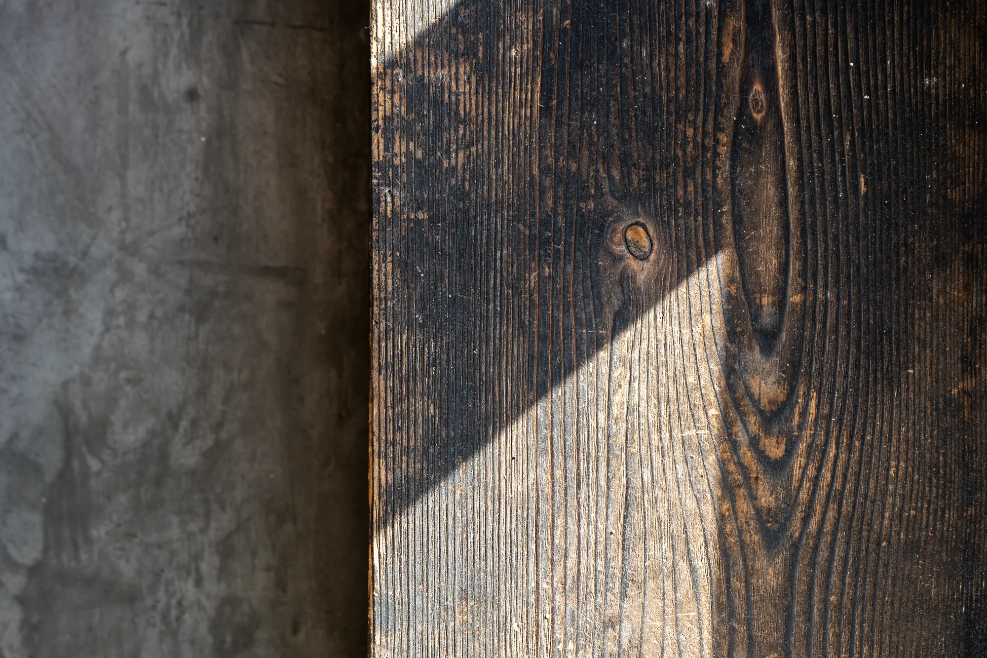 Detail of charred timber beside raw concrete, light cutting across the burnt grain