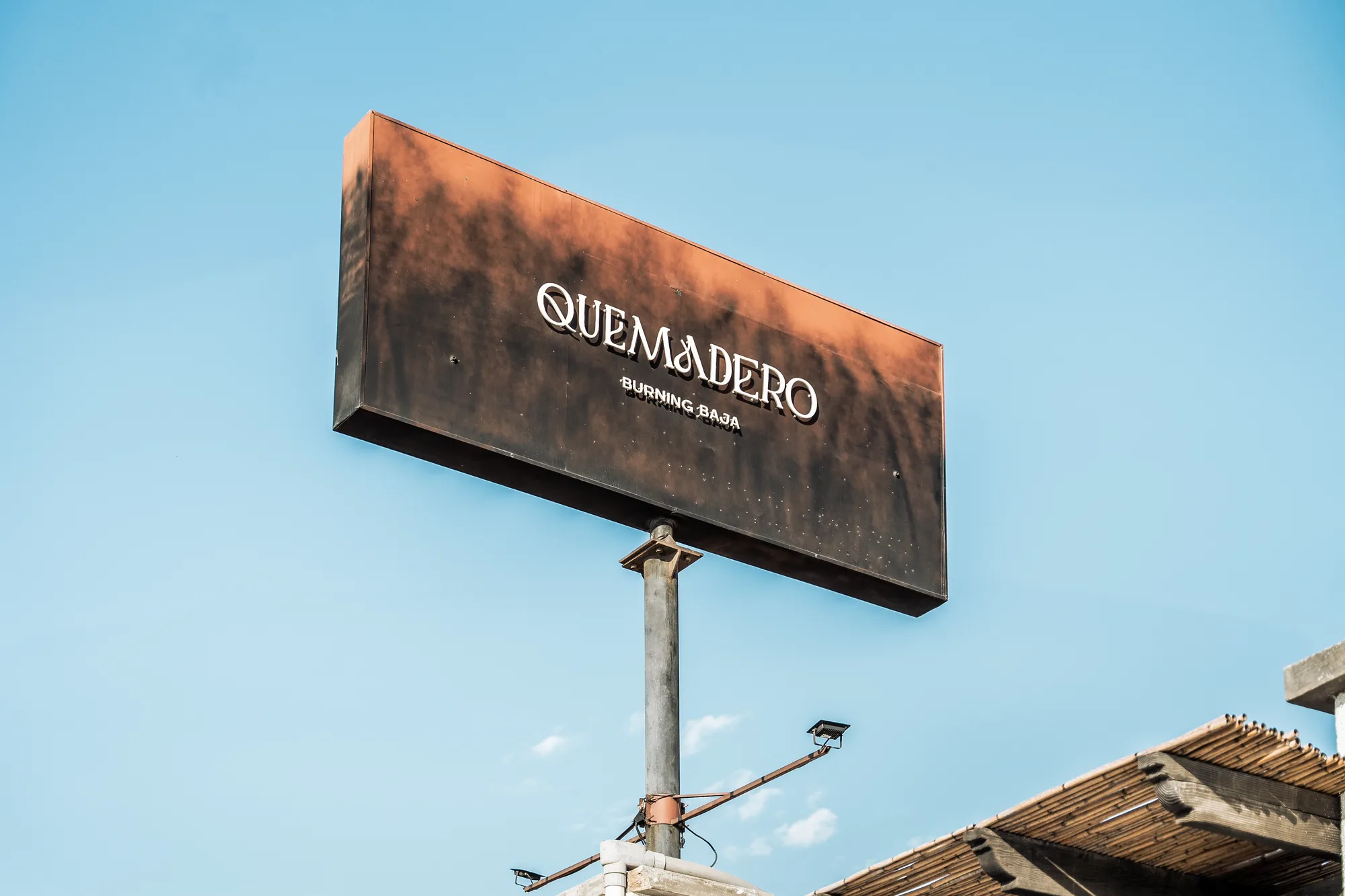 The rusted steel Quemadero sign, reading Burning Baja, against a clear sky above the reed roof