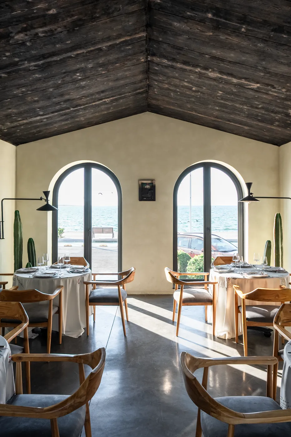 Dining room under a dark timber ceiling with two arched openings to the sea, set tables and wood chairs on polished concrete