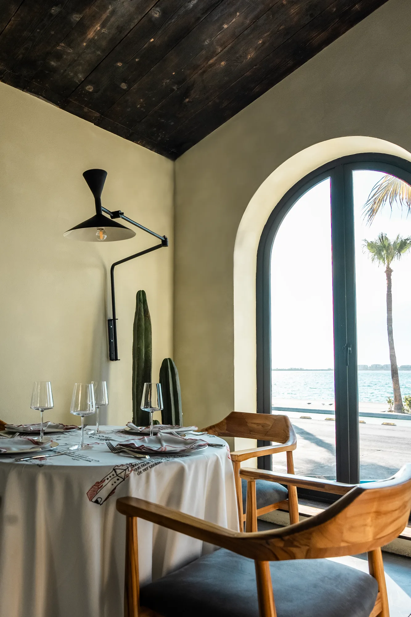 Corner table set with wine glasses by an arched window framing a palm and the Sea of Cortez, an articulated black wall lamp above