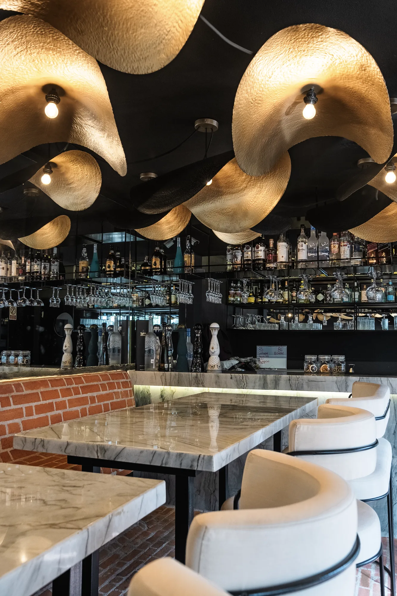 Bar under a ceiling of wavy gold pendants, marble tables with white chairs and a bottle-lined back bar over brick
