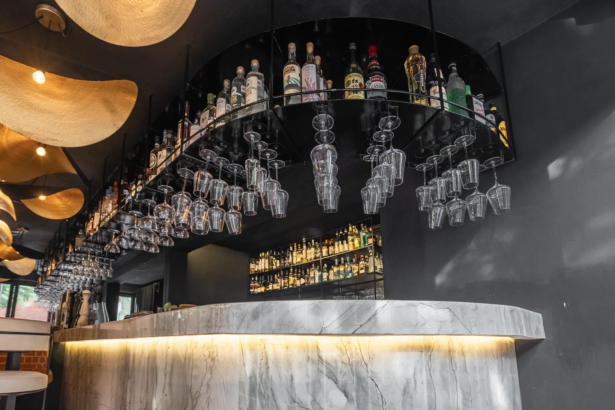 Curved marble bar with a backlit base, a suspended rack of inverted glasses and liquor shelves under wavy gold pendants