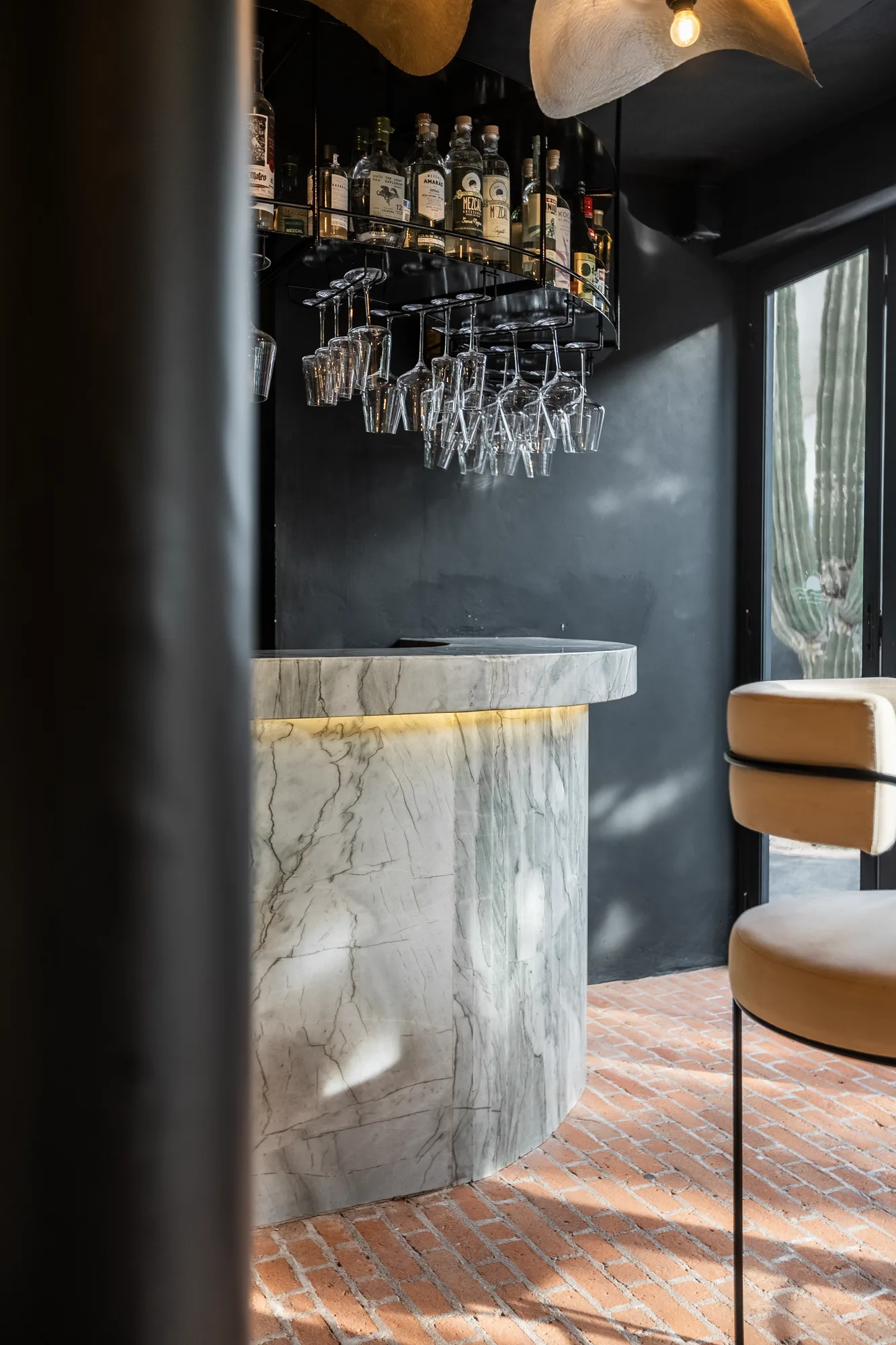 Detail of the curved marble bar with a brass-lit base and hanging glasses, a cactus glimpsed through the window on a brick floor
