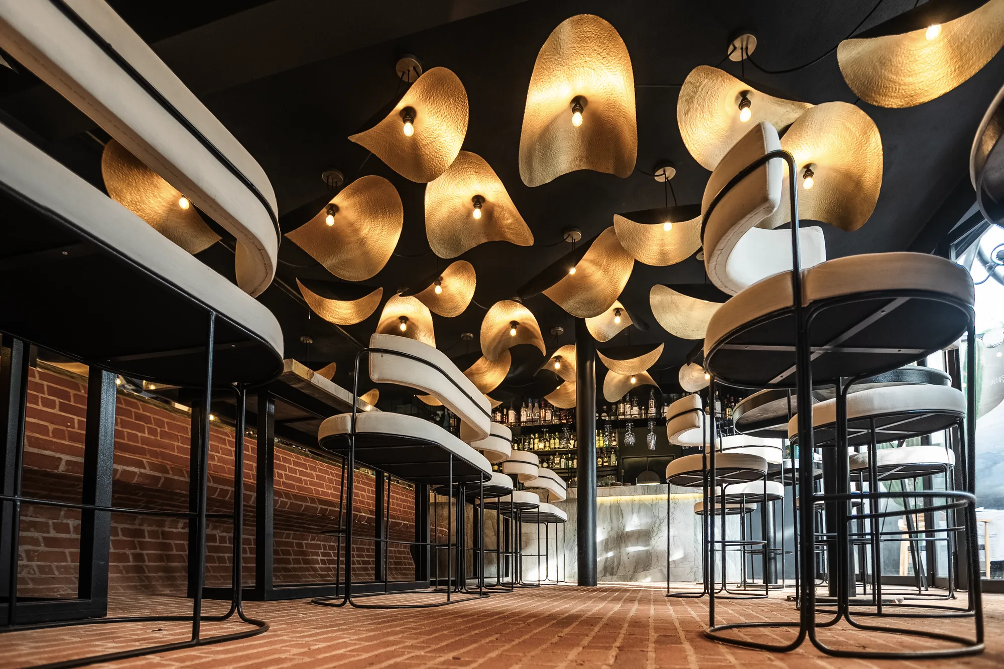 Bar room beneath a sculptural ceiling of wavy gold discs, upholstered stools at the counters and brick walls and floor