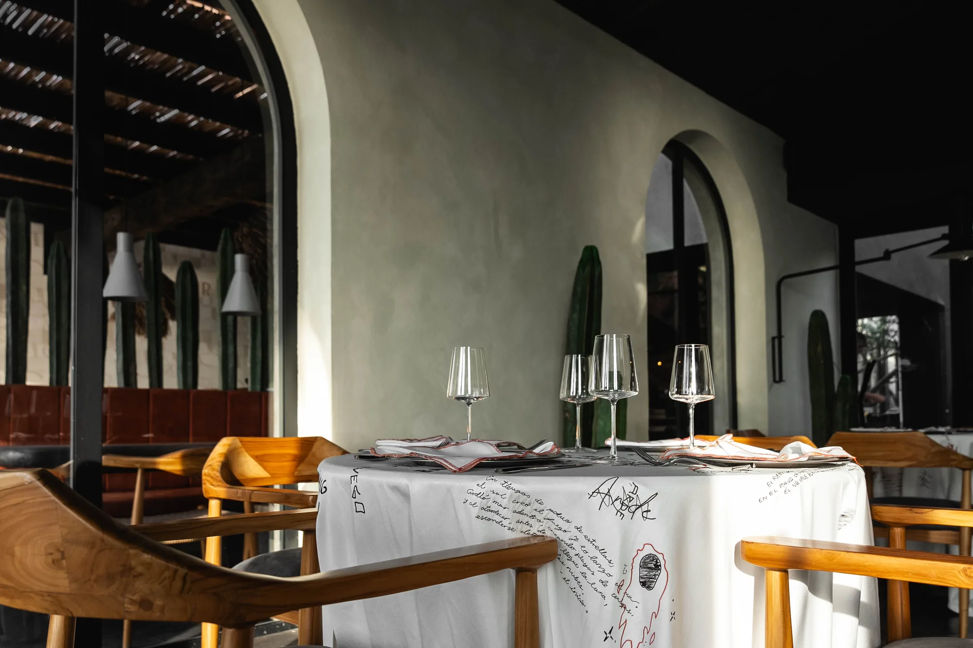 Table set with wine glasses on the signature poem-printed linen, teak chairs and cacti under a reed roof by arched openings