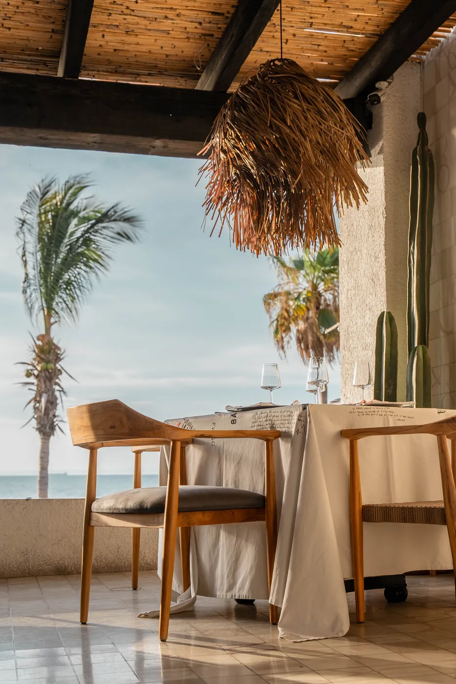 Terrace table under a woven palm-frond pendant with teak chairs, opening to palms and the Sea of Cortez, Quemadero in La Paz