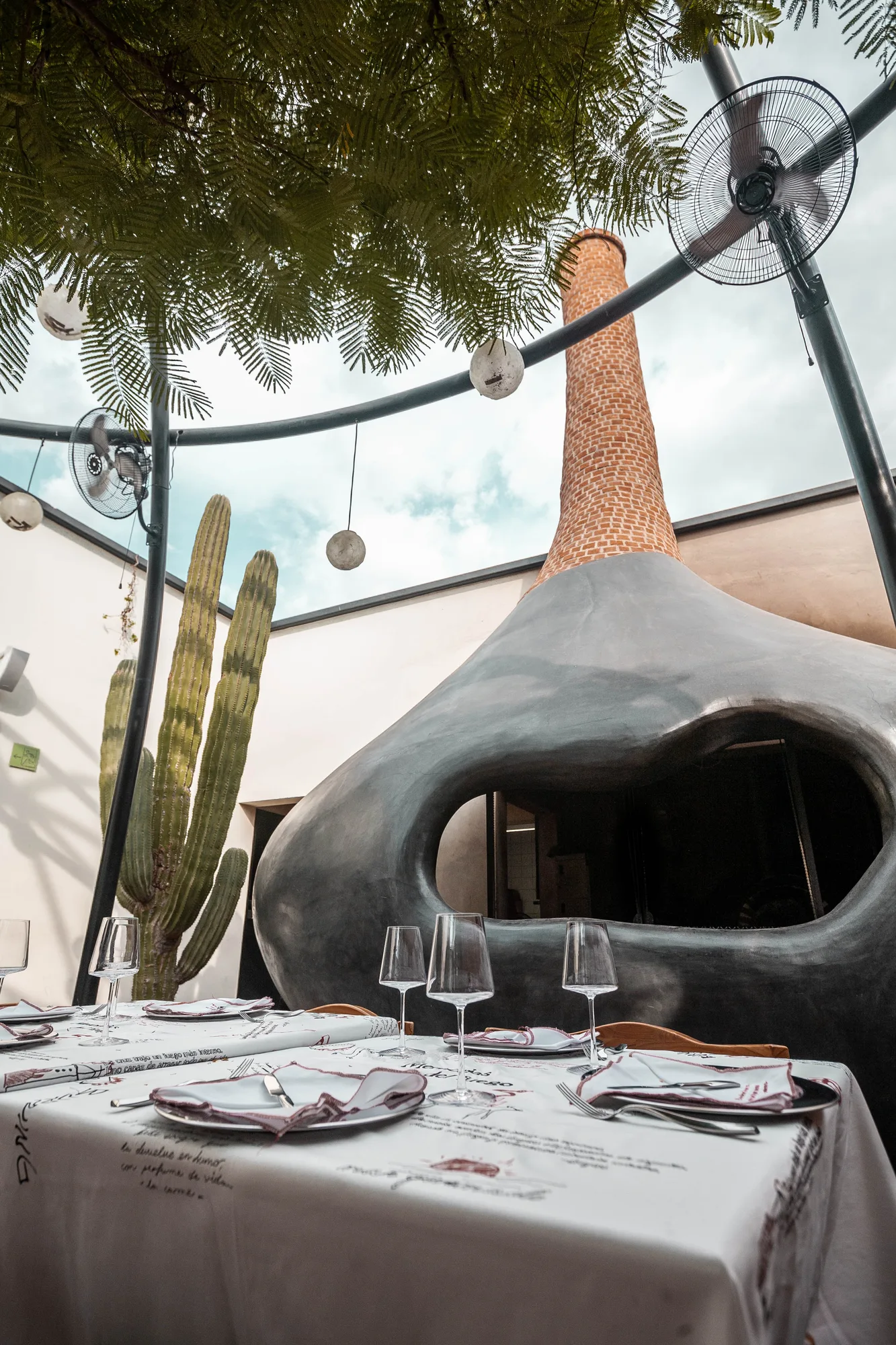 Open courtyard with the black sculptural wood-fired oven and its brick chimney rising among cacti and misting fans, a table set below