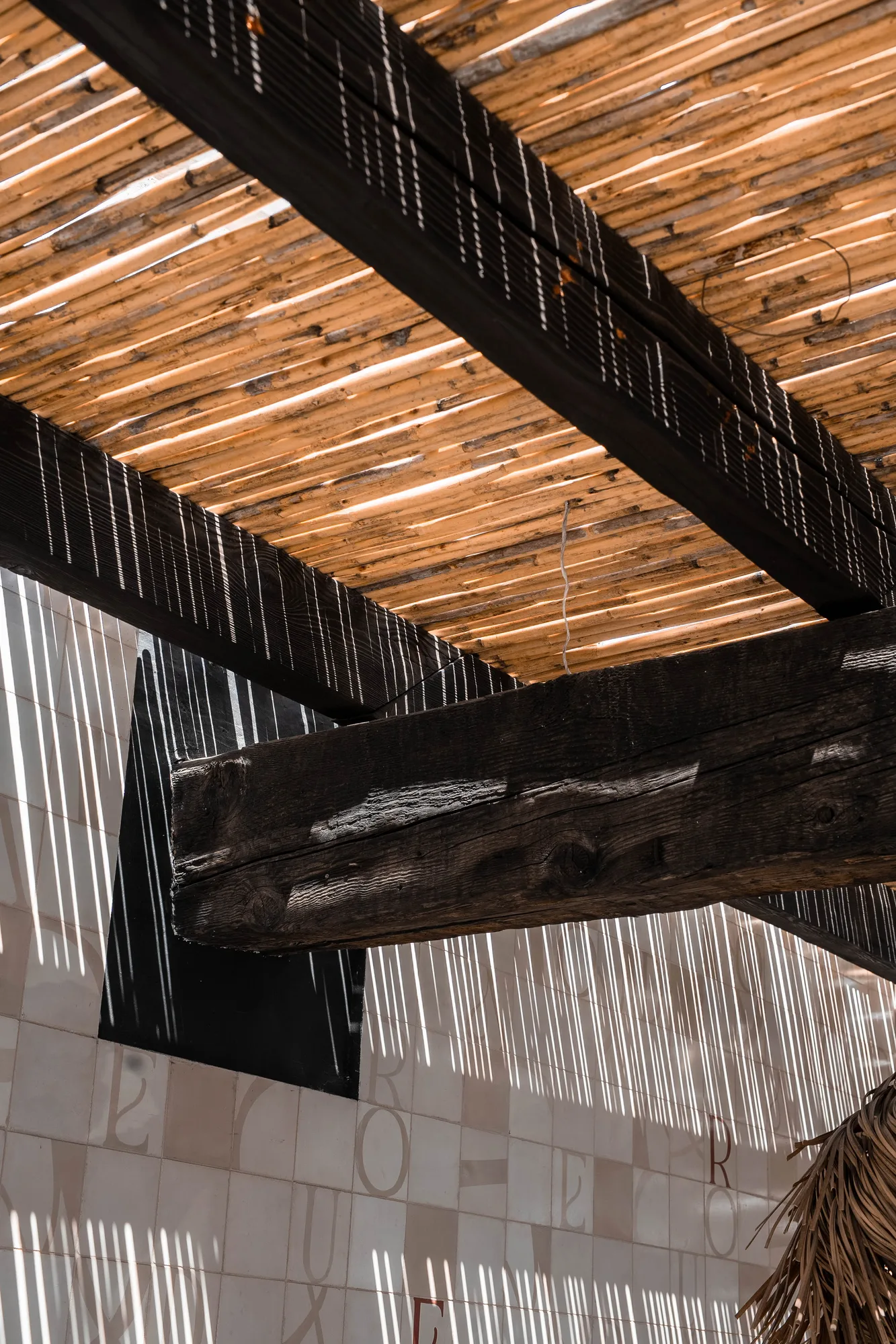 Looking up at charred timber beams supporting a reed ceiling, light filtering through onto a lettered tile wall