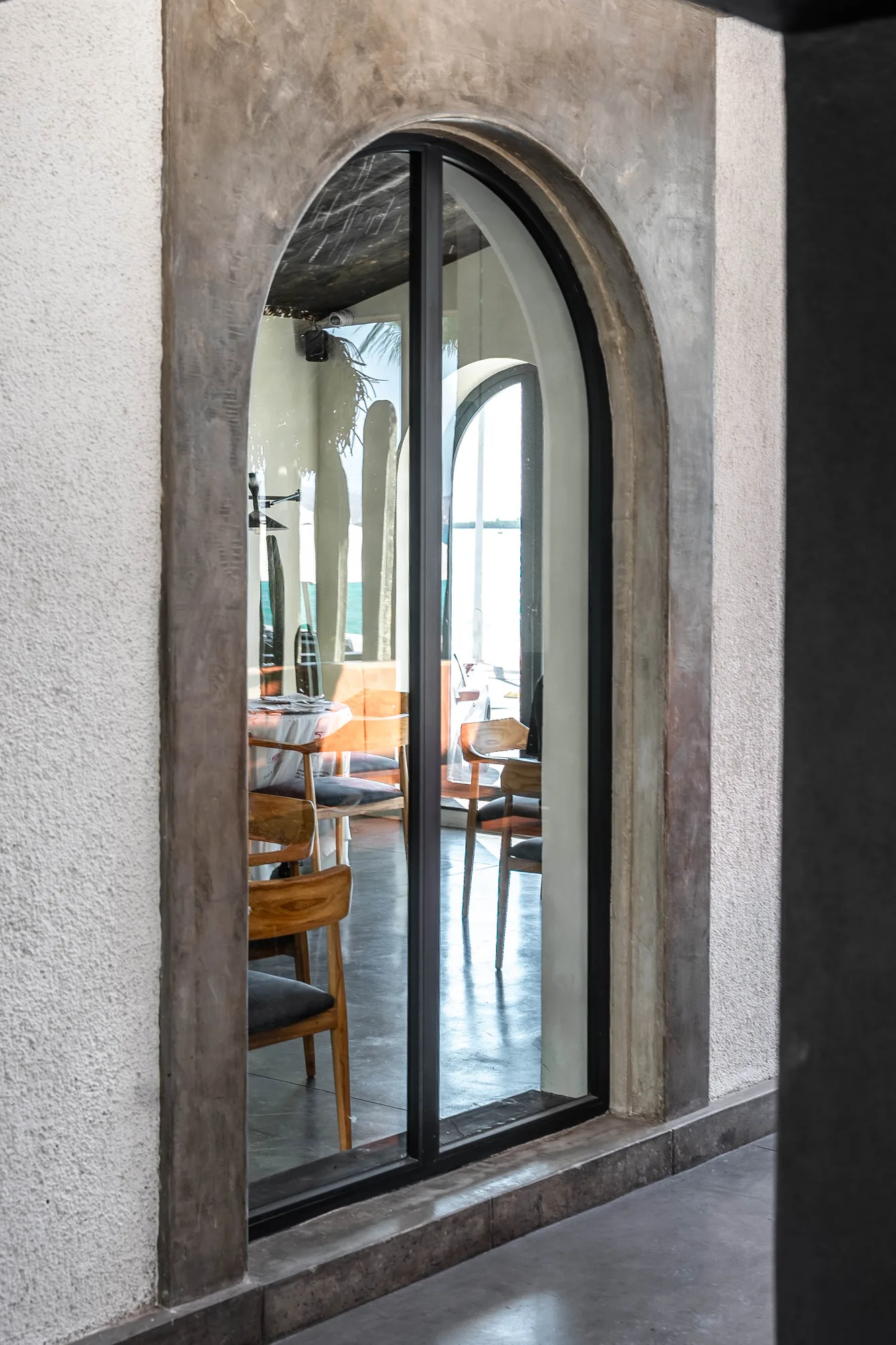 View through a black-framed arched window into the dining room with tables and wood chairs on polished concrete